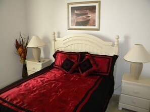 A bedroom with a white bed, red and black bedding, two bedside lamps, a nightstand, a vase with dried plants, and a framed picture on the wall. - Ip60166 - Highgate at Legacy Park - 4 Bed 3 Baths Villa (Davenport)