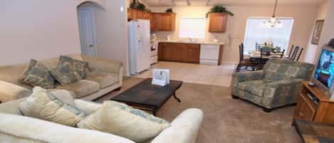 A living room with a sofa, armchair, and coffee table. There is a kitchen in the background with wooden cabinets and a refrigerator.