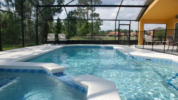 A swimming pool with a glass enclosure, surrounded by a deck with chairs and a view of trees and houses.