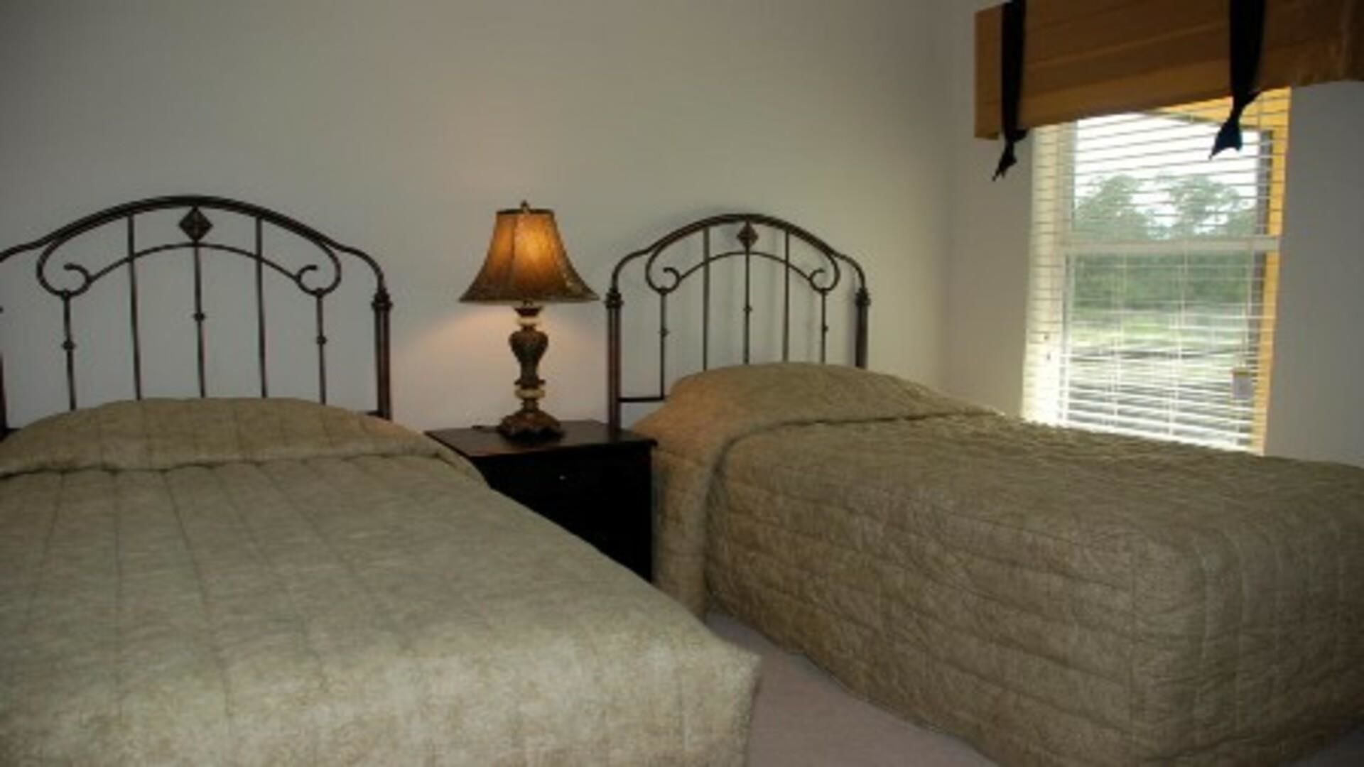 A bedroom with two beds, a nightstand with a lamp, and a window with blinds.