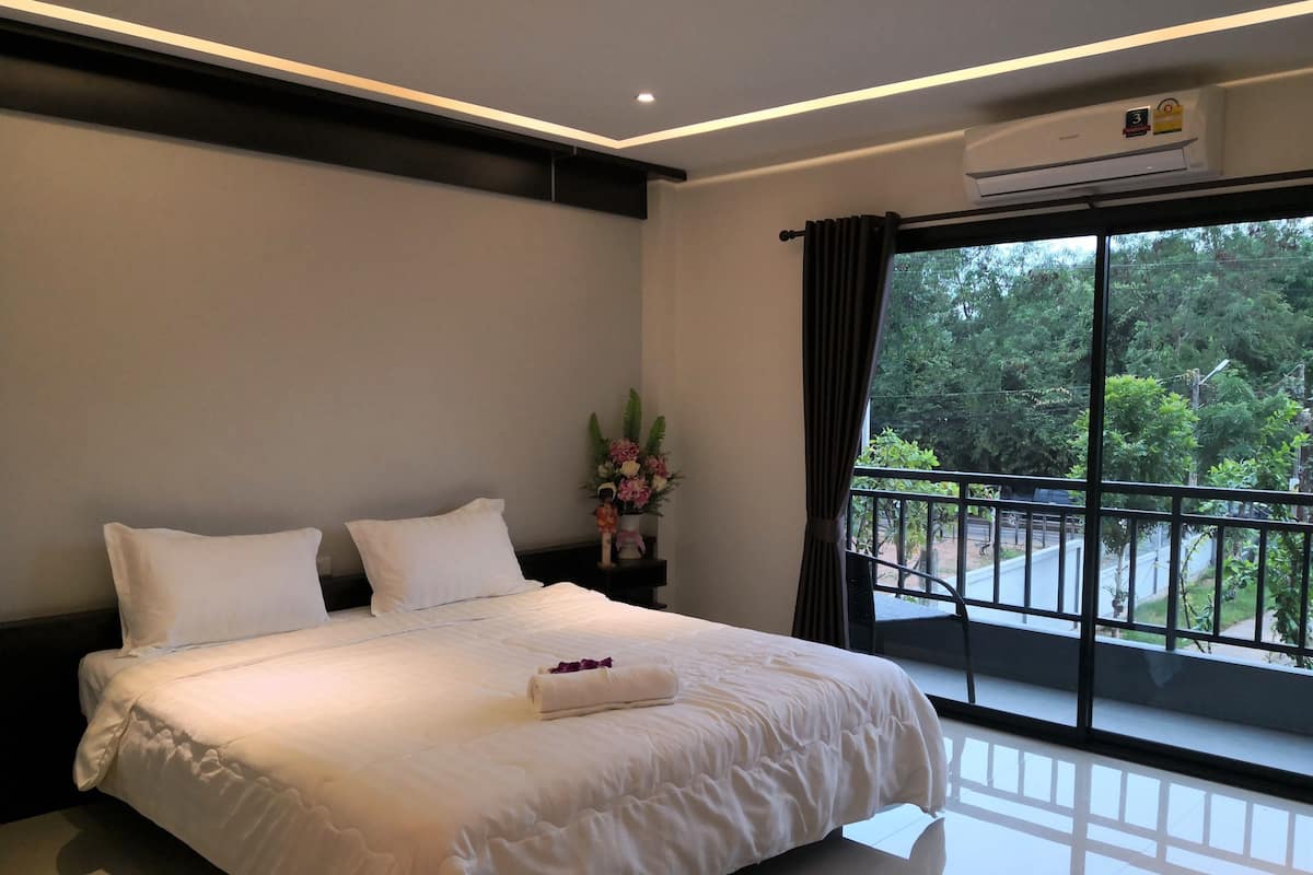 Une chambre d’hôtel avec un grand lit, une table de chevet ornée d’un vase de fleurs et un balcon donnant sur un paysage verdoyant.