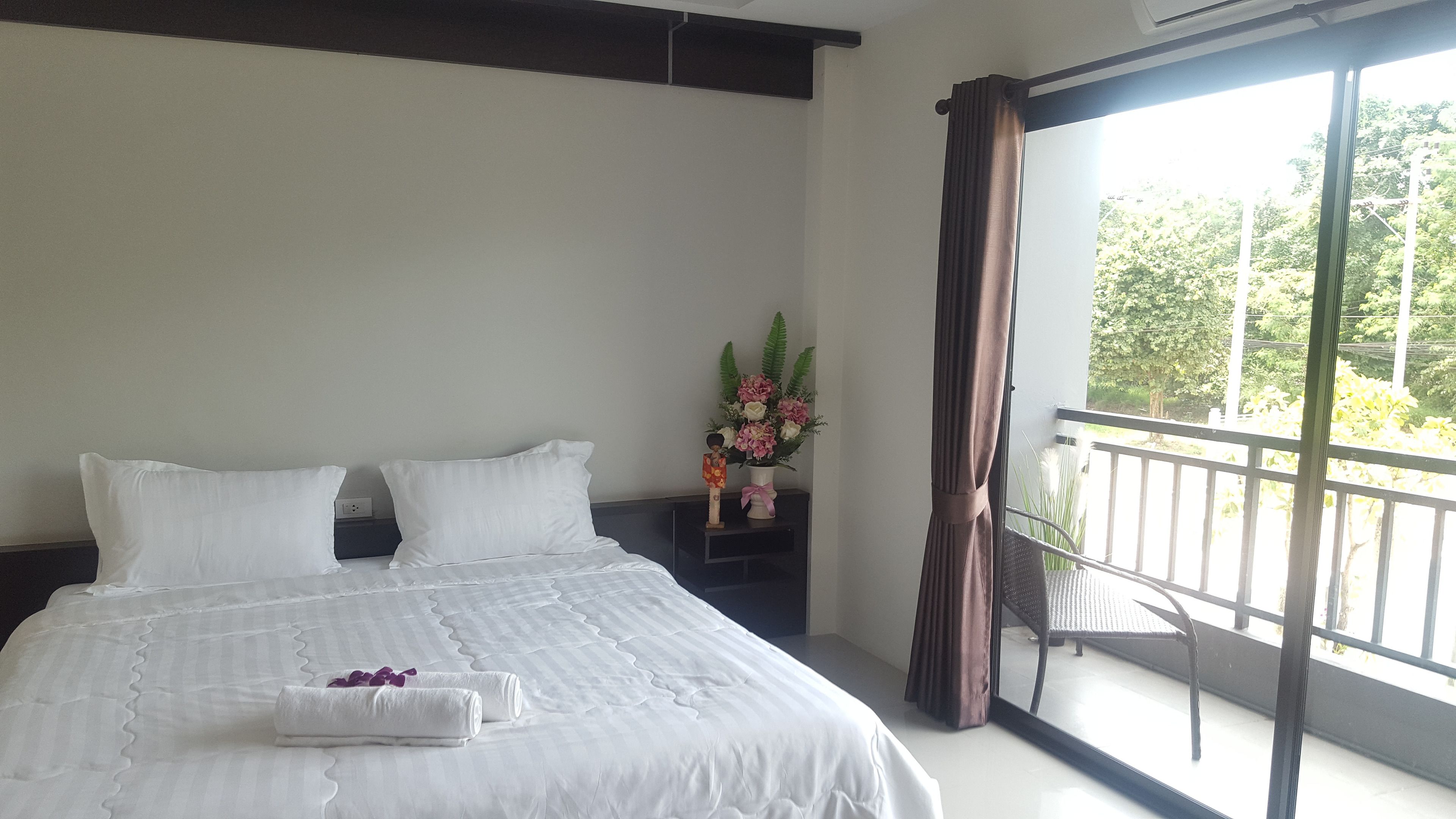 Habitación de hotel con una cama grande, una mesita de noche con un florero y vistas a un balcón con follaje.