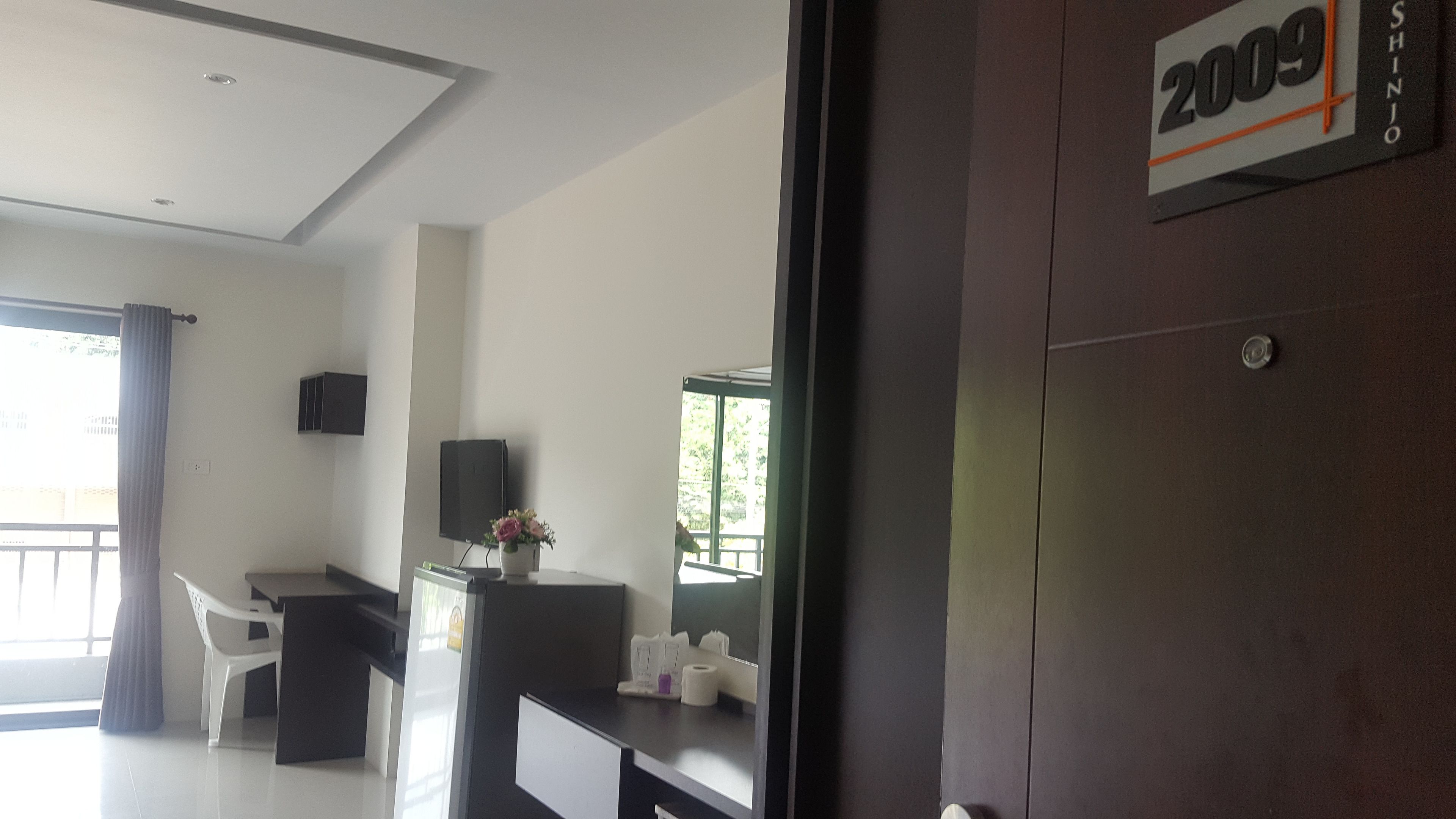 A modern hotel room with a large window, a flat-screen TV, a desk with a computer, and a small table with a vase of flowers.