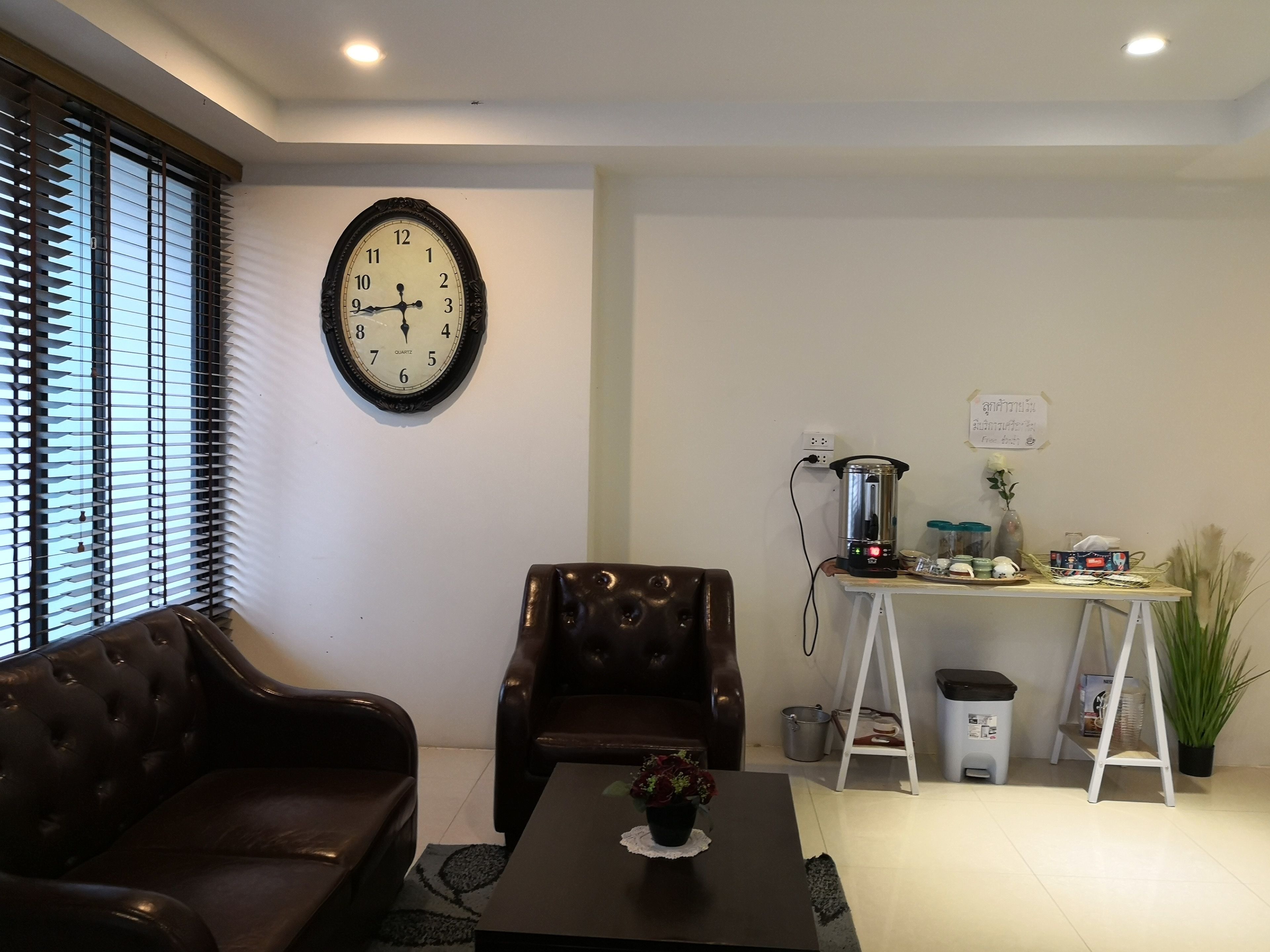 A living room with a brown leather sofa, a coffee table with a vase, a clock on the wall, and a small table with a kettle and other items.