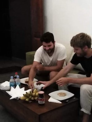 Two men sitting on a couch with a table in front of them. The table has food and drinks on it.