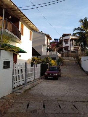 A narrow street with a white picket fence, a parked car, and houses with balconies and a sloped roof.