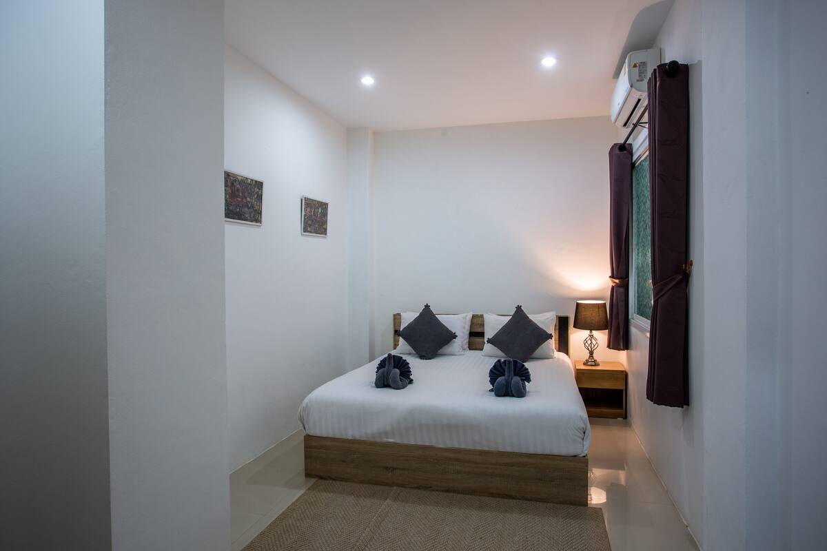 A bedroom with a wooden bed frame, white bedding, two pillows, a bedside table with a lamp, and a wall-mounted air conditioner.