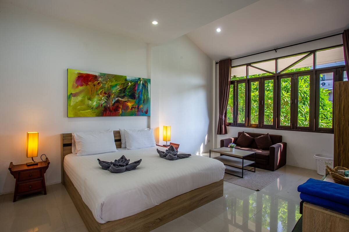 A modern hotel room with a large bed, a painting on the wall, a sofa, and a view of greenery through the windows.