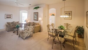 Un salon comprenant un canapé, un fauteuil, une table basse et une table à manger avec des chaises. On y trouve également un ventilateur de plafond, une lampe et un lustre.