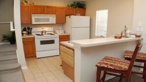 Une cuisine compacte avec des meubles en bois, une cuisinière blanche, un four à micro-ondes et un réfrigérateur. On y trouve également un petit îlot avec deux chaises.