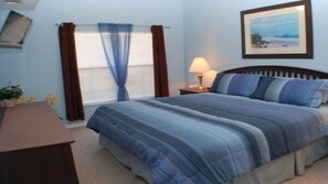 Une chambre à coucher avec un lit, une table de chevet, une lampe, une fenêtre avec des rideaux et un tableau encadré au mur.