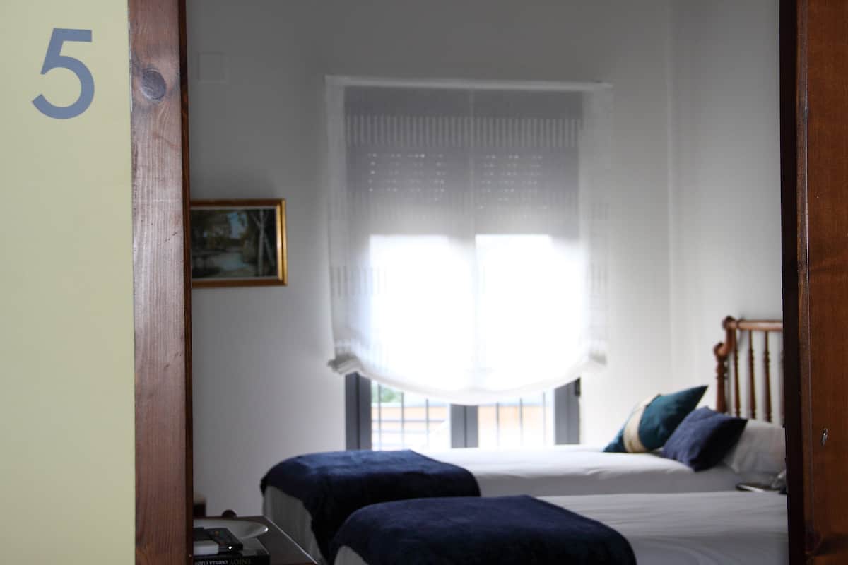 A room with two beds, a window with sheer curtains, a framed picture on the wall, and a door with a number '5' on the wall.