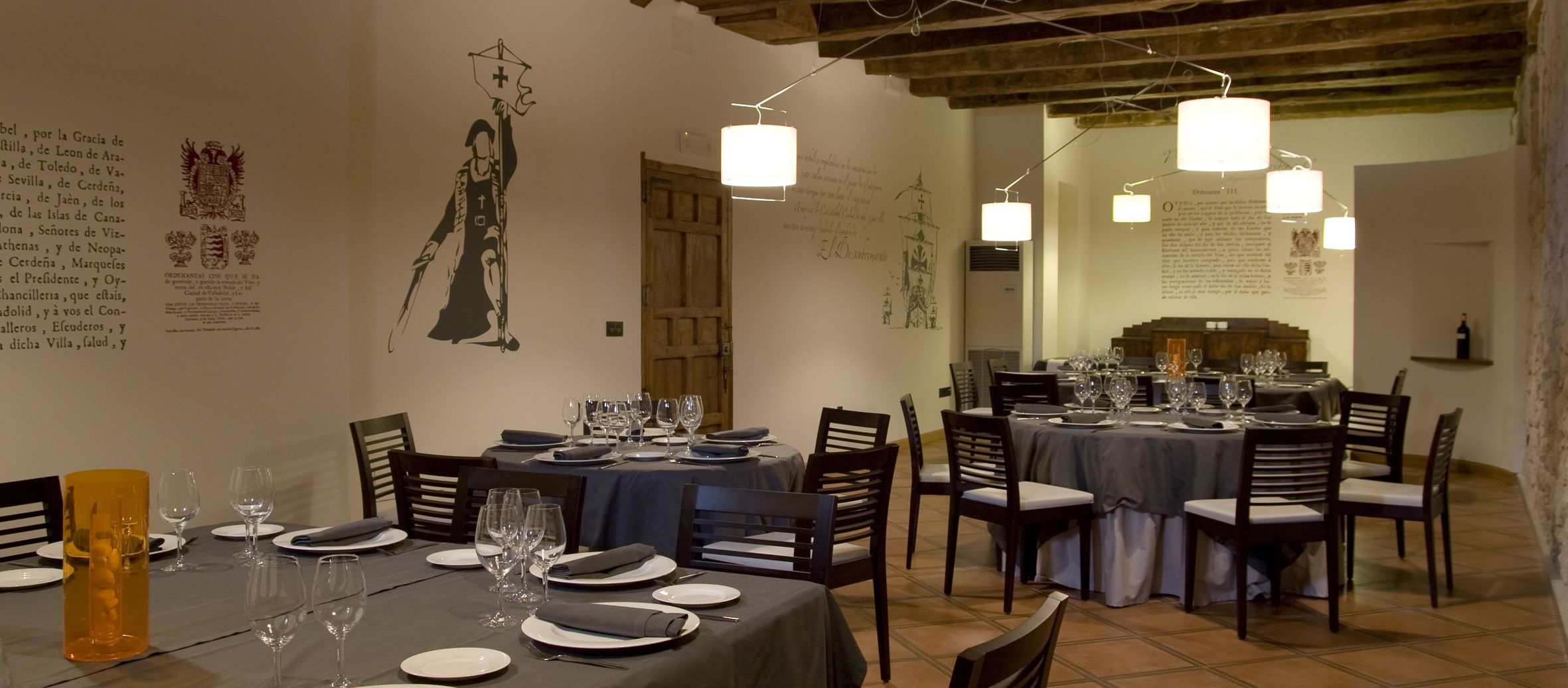 A dining area with tables set for a meal, featuring a traditional European architectural style with exposed wooden beams and tiled flooring.