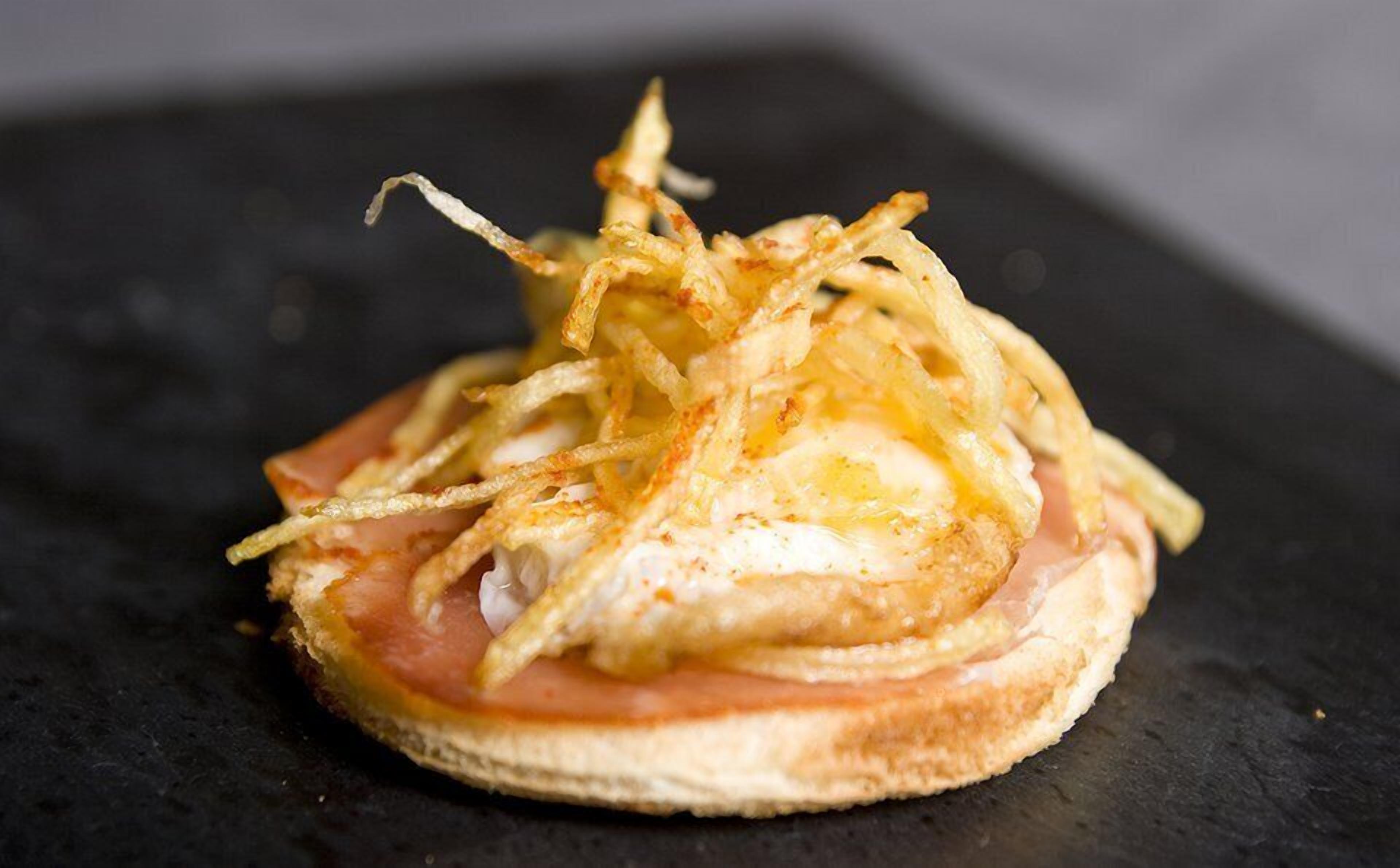 Un sándwich con rebanada de pan, cubierto con jamón, un huevo frito y cebollas fritas crujientes.