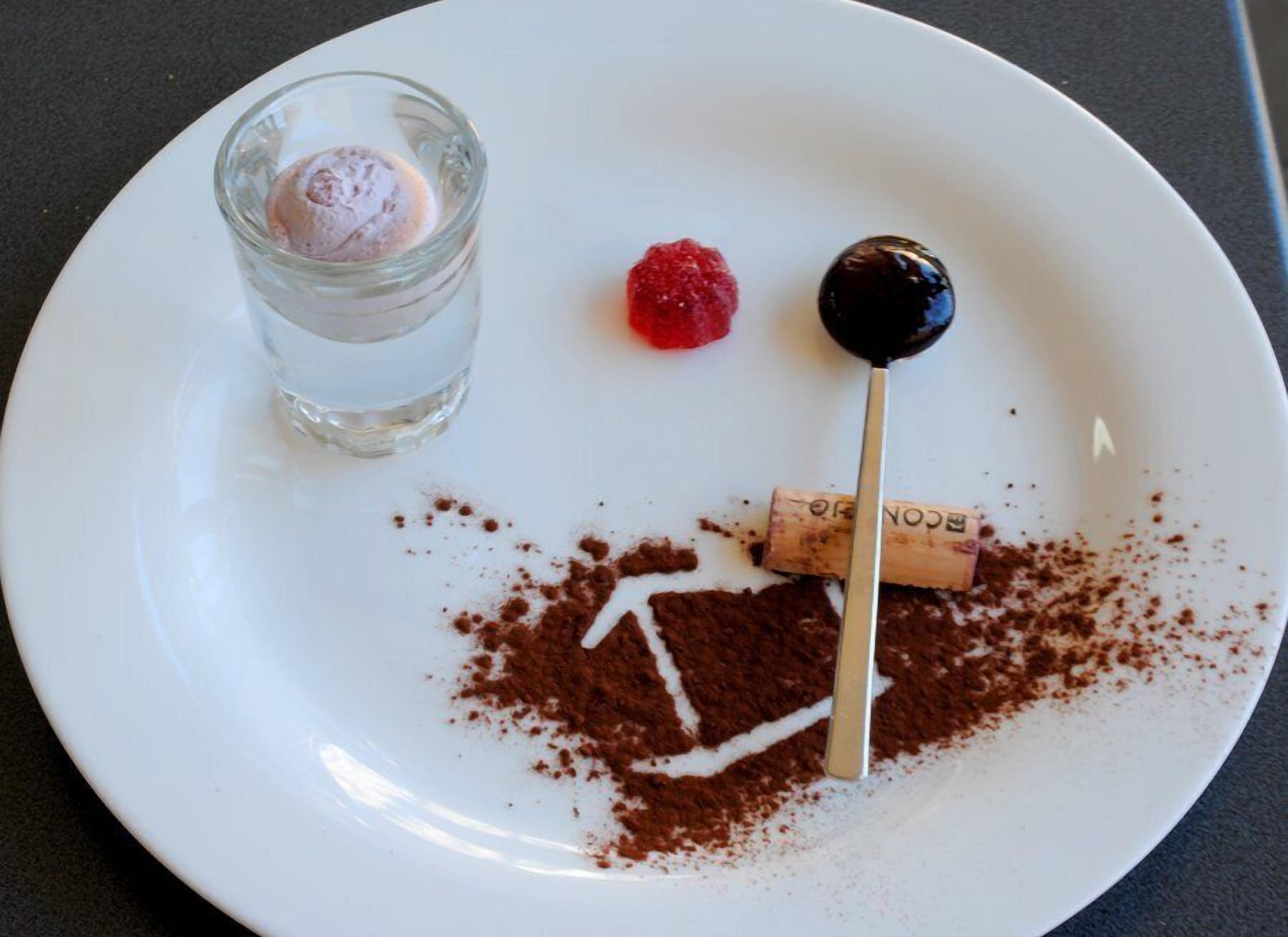 Un plat blanc amb un postre que consisteix en una bola de gelat, una frambuesa i una cirera amb cobertura de xocolata en un pal.