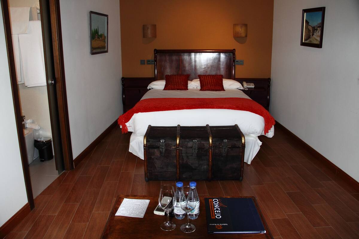 A hotel room with a large bed, a wooden headboard, a red throw blanket, a nightstand with a phone, and a suitcase on the floor.