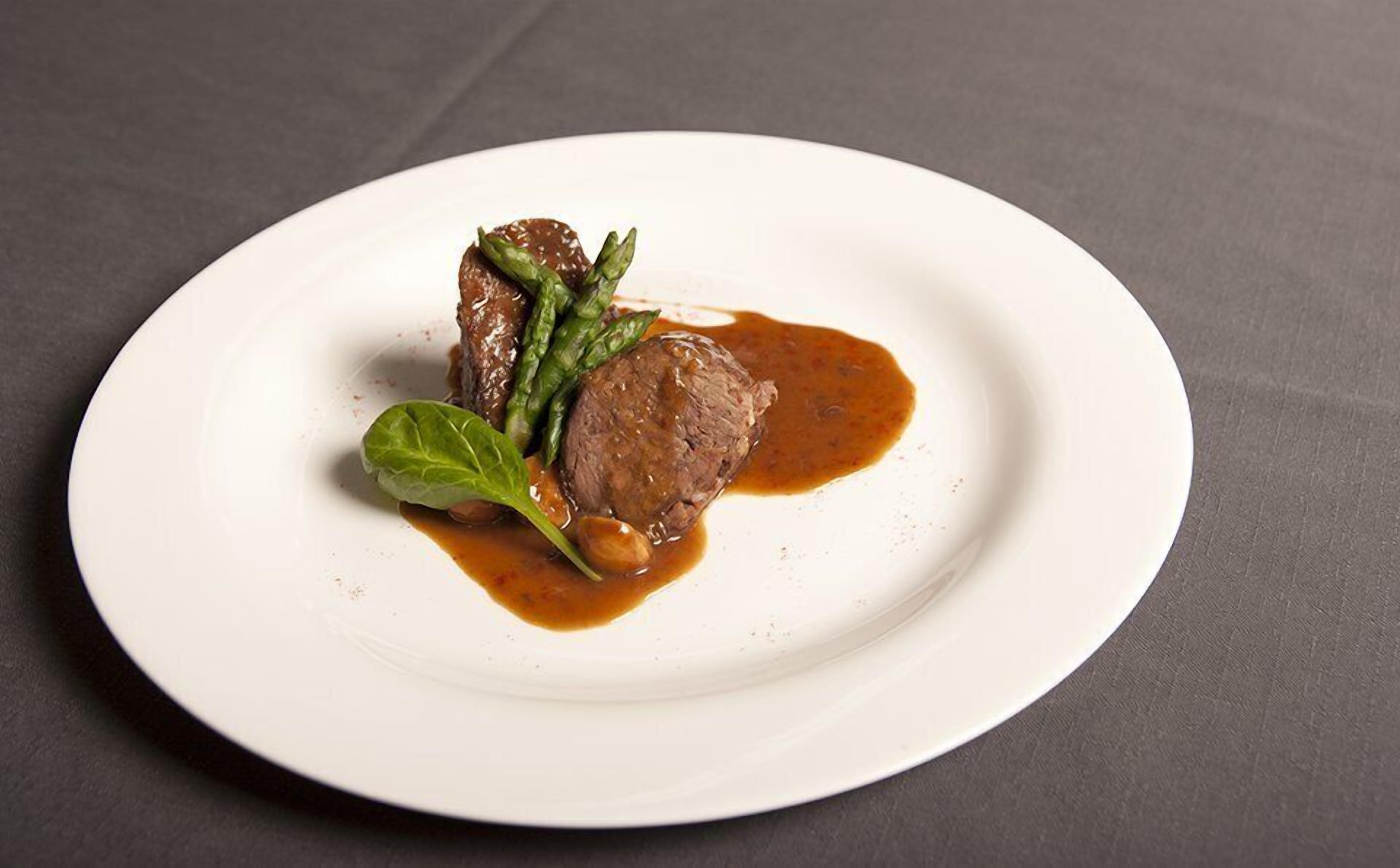 A white plate with a steak, asparagus, and a sauce on a dark surface.