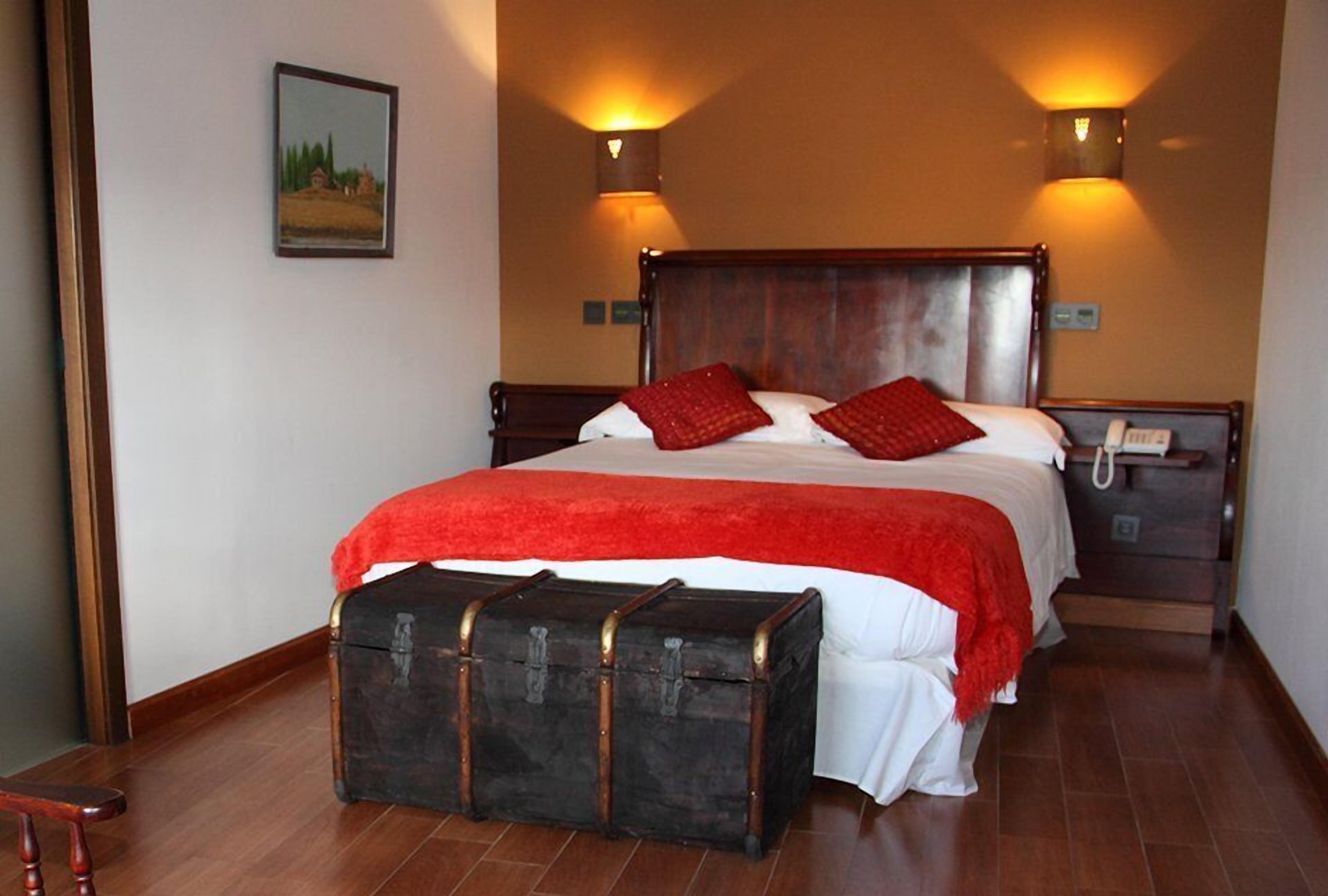 A bedroom with a wooden bed, red pillows, a red blanket, a wooden chest of drawers, and two wall-mounted lamps.