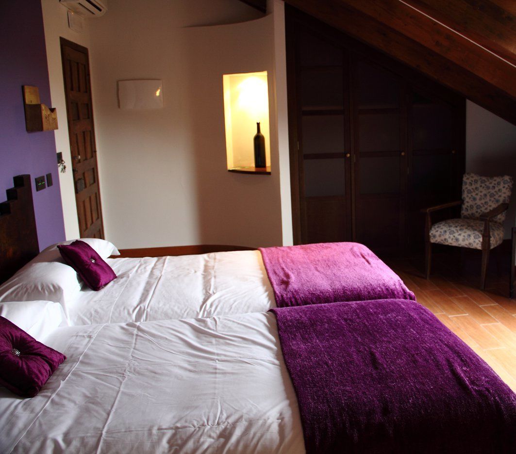 Una cama con ropa de cama blanca y morada, una silla de madera y una lámpara fijada a la pared.