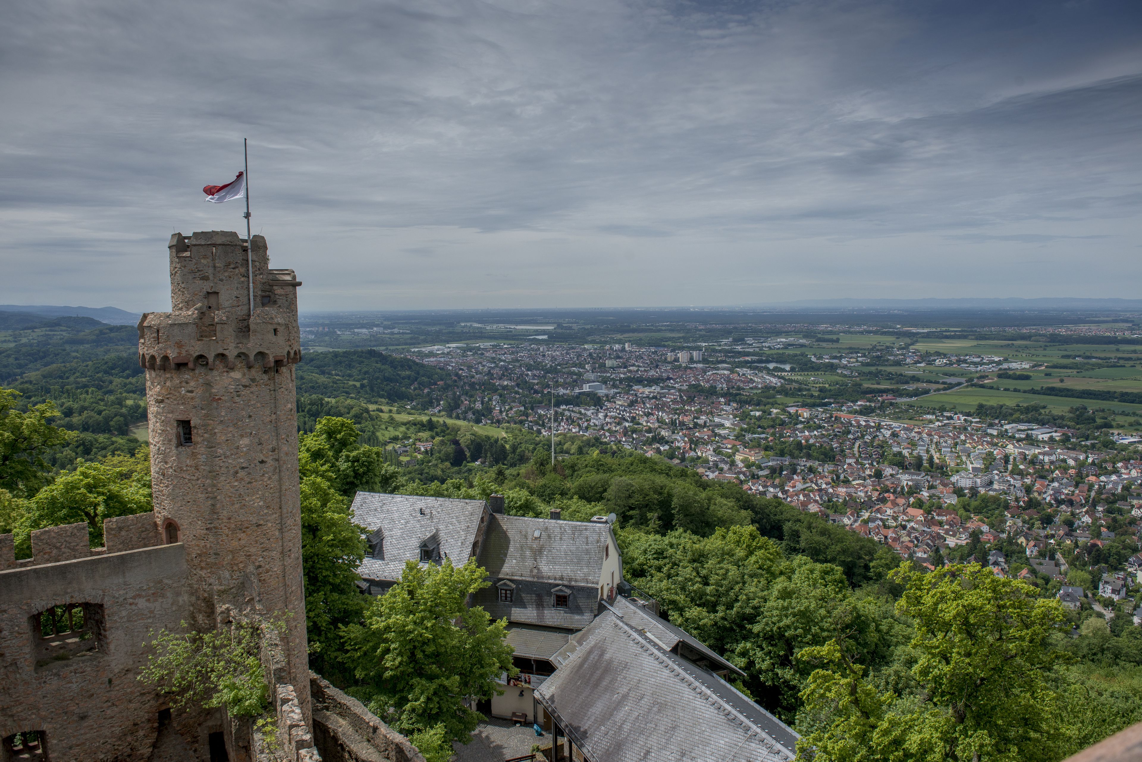 A historic stone tower with a flag, surrounded by lush greenery and a view of a town below.