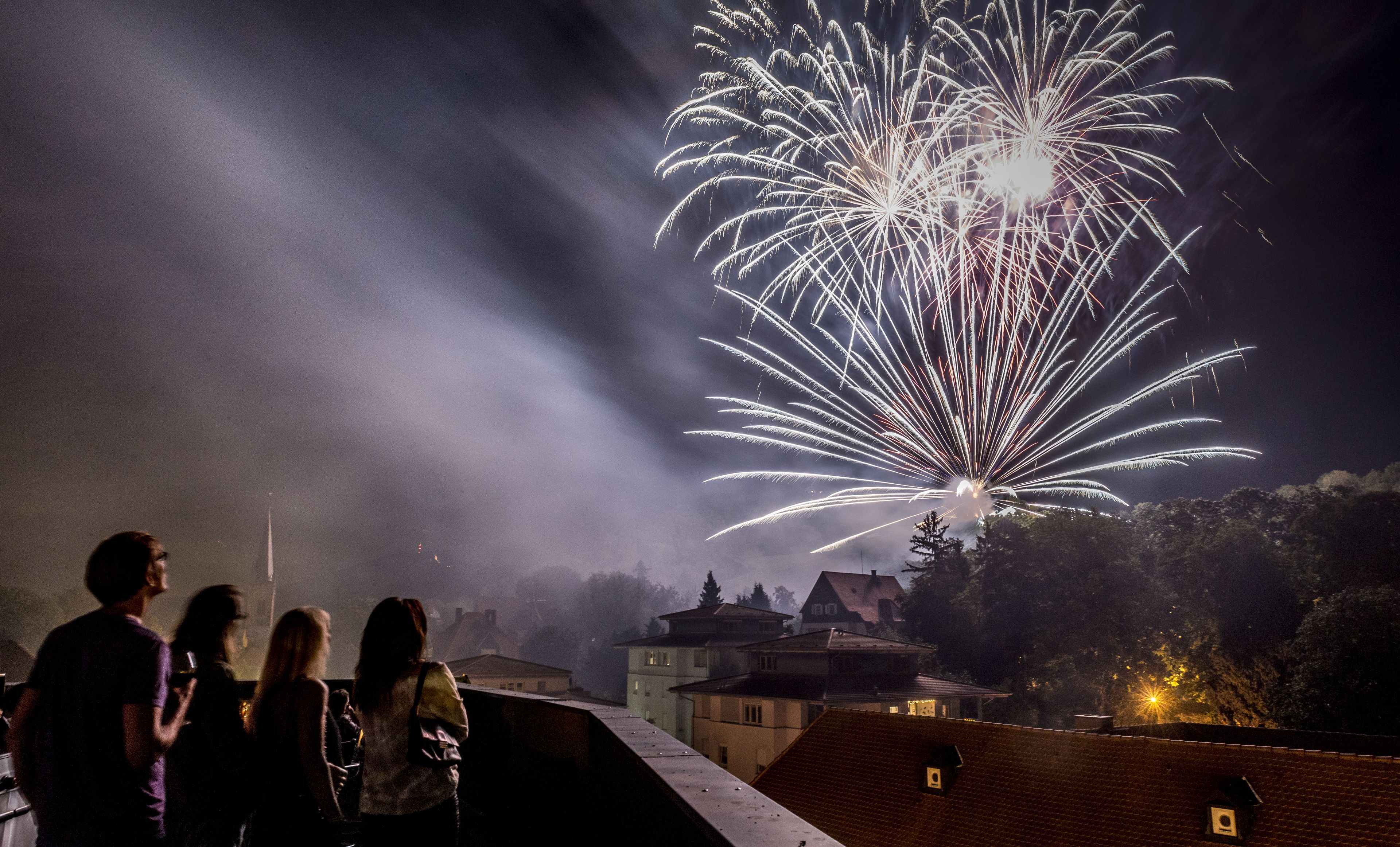 Cena noturna com fogos de artifício, um grupo de pessoas em uma varanda e edifícios ao fundo.