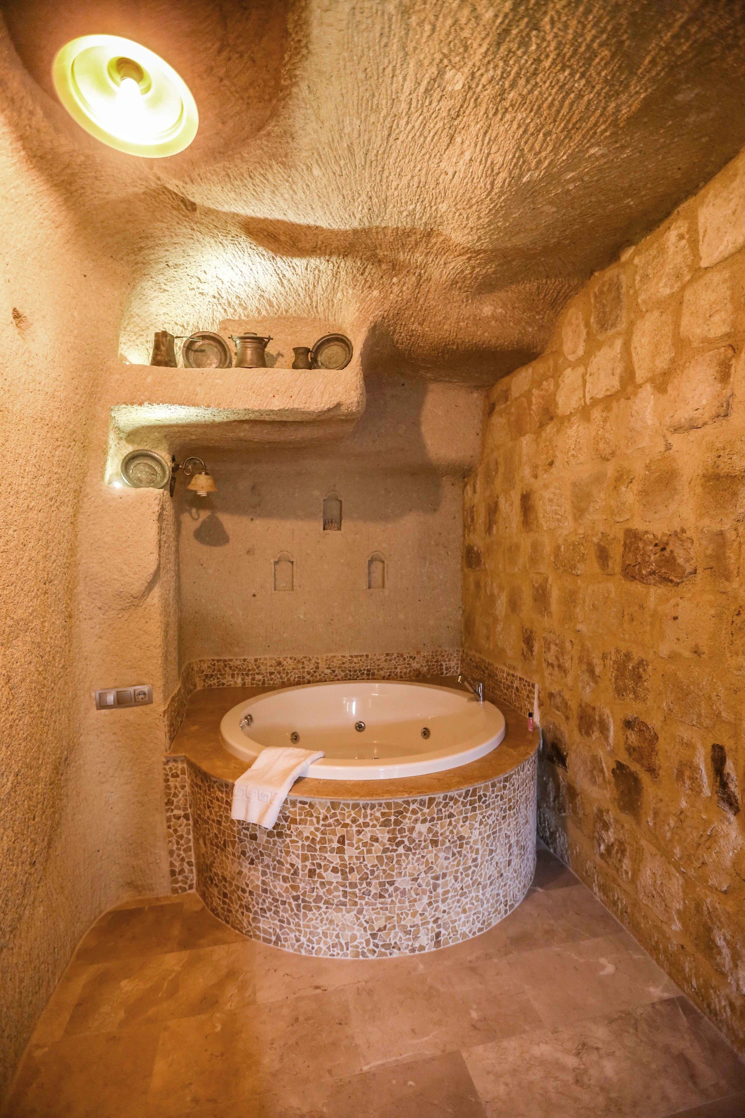 A stone-walled bathroom with a mosaic-tiled bathtub, a towel on the edge, and a light fixture above.