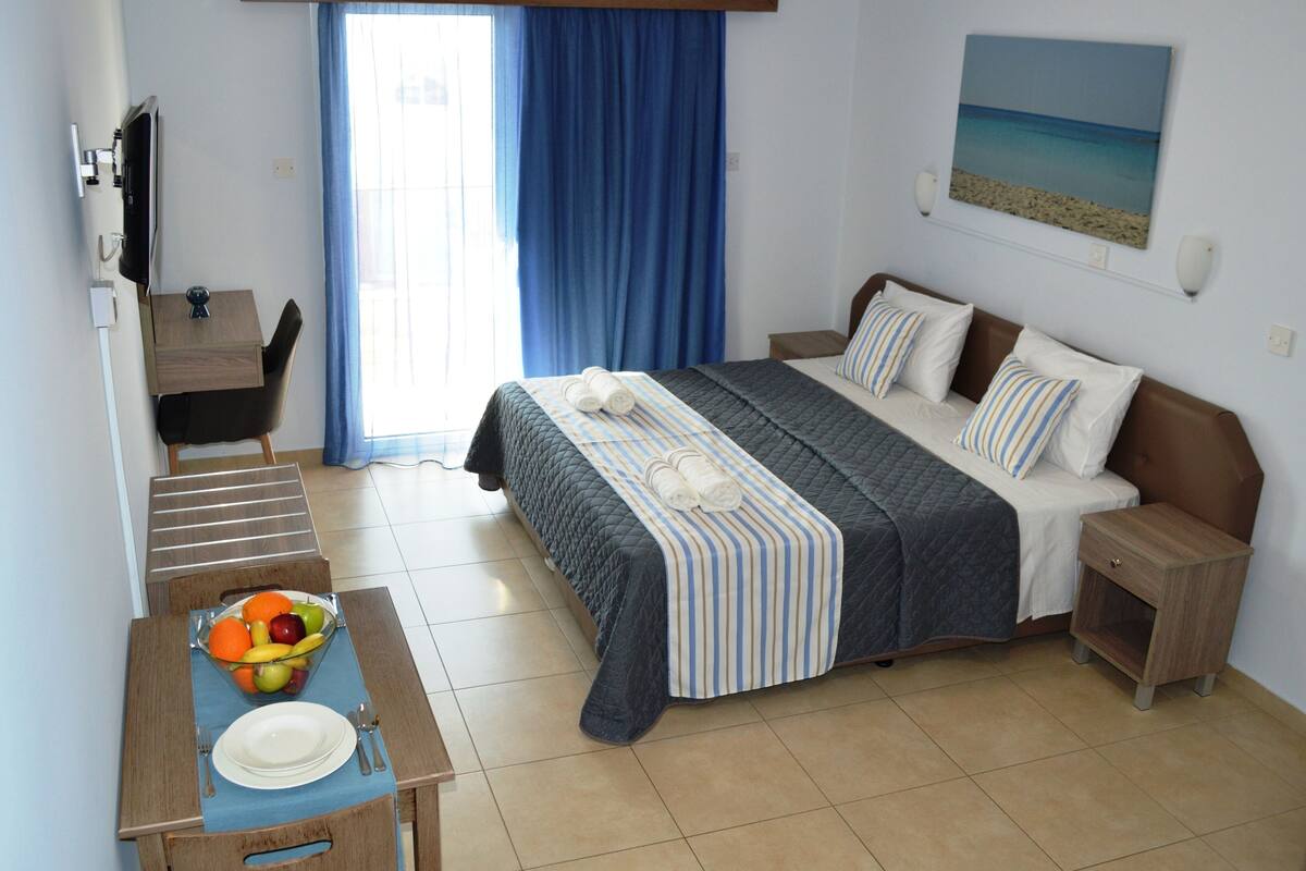Une chambre d’hôtel avec un lit, un bureau sur lequel se trouve un panier de fruits, une chaise, un téléviseur et un tableau accroché au mur.