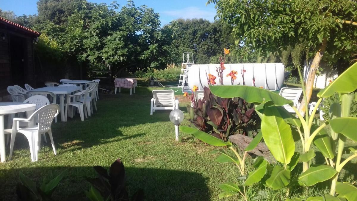 Un área de patio al aire libre con mesas y sillas de plástico blanco, rodeada de abundante follaje y un tanque de agua blanco al fondo.