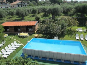 Una piscina rectangular con revestimiento azul, rodeada de sillas reclinables blancas y un edificio de madera con techo de tejas rojas.