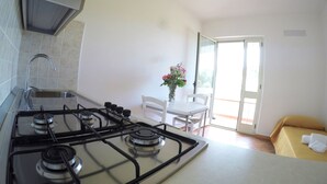 Une cuisine équipée d’une cuisinière à gaz, d’un évier et de murs recouverts de carreaux blancs. On y trouve une table à manger avec des chaises et un vase rempli de fleurs. On aperçoit, au fond, un lit recouvert d’une couverture jaune.