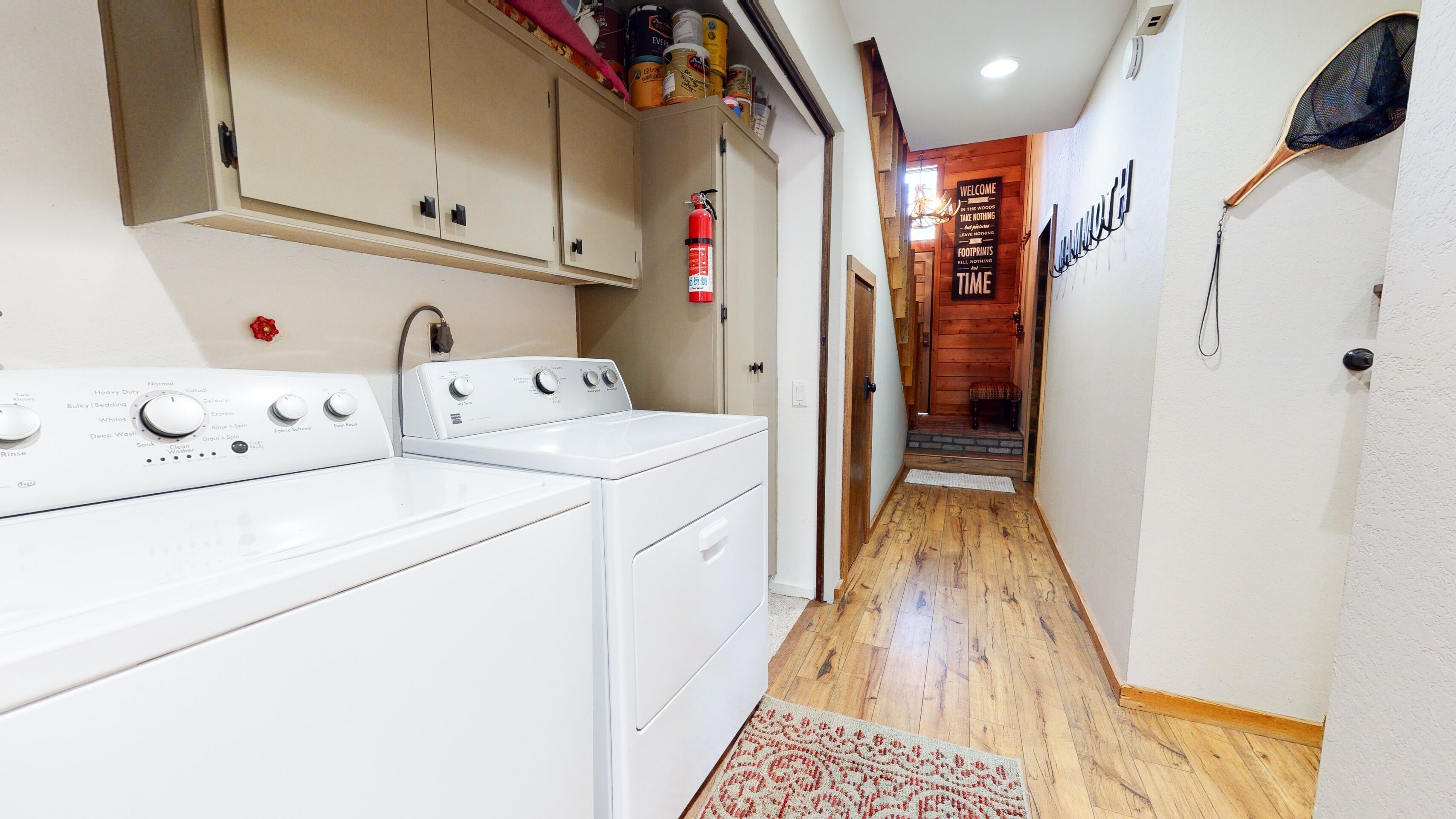 Een wasruimte met een wasmachine en droger, een brandblusser en een gang die leidt naar een houten kamer.
