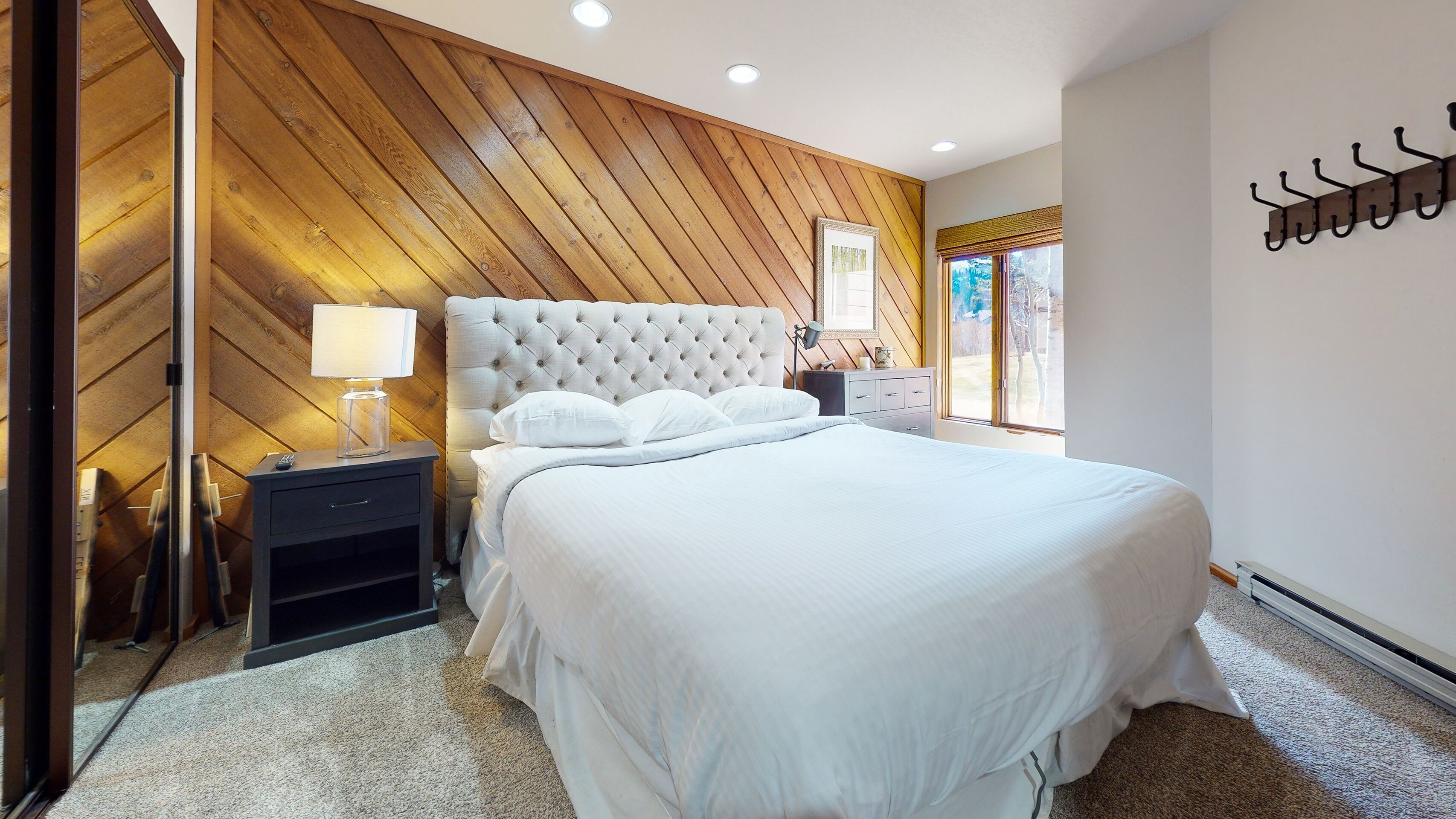 A bedroom with a large bed, wooden panel walls, a nightstand with a lamp, and a window with a view of the outdoors.