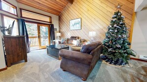 Ein gemütliches Wohnzimmer mit einem braunen Ledersofa, einem Couchtisch und einem Weihnachtsbaum. Die Decke ist aus Holz und es gibt ein großes Fenster mit Vorhängen.