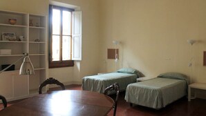 A room with two beds, a wooden table, a chair, a desk, a lamp, and a shelf with various items.