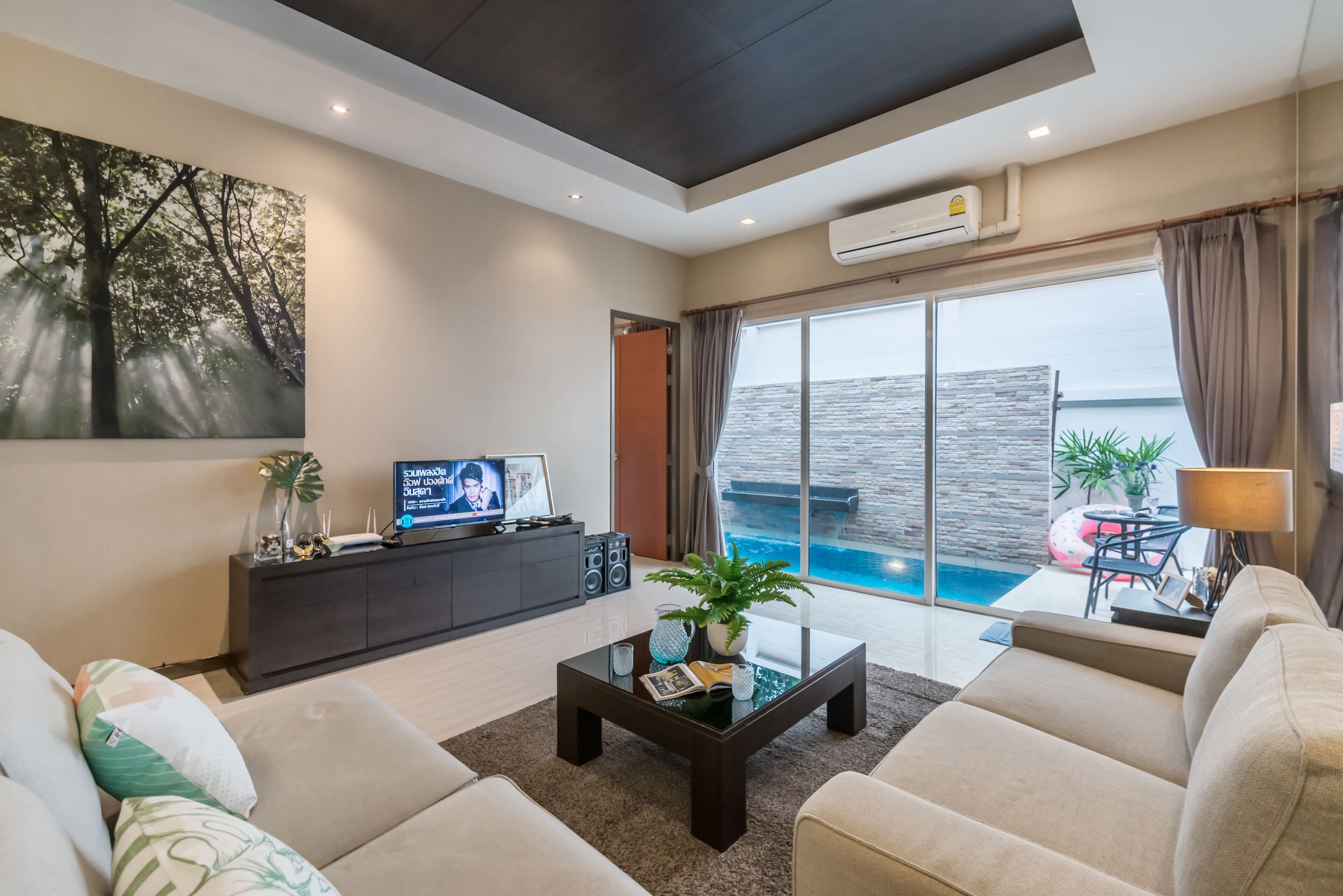 A modern living room with a large window overlooking a pool, a flat-screen TV, and a dark coffee table with decorative items.