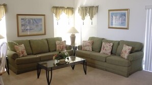 A living room with a green sofa, a glass coffee table, and floral cushions. There are two framed pictures on the walls. - Ahr123 - Hampton Lakes - 5 Bed 3 Baths Villa (Davenport)