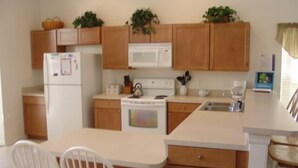 A compact kitchen with wooden cabinets, a microwave, a stove, and a sink. There are two white chairs and a small table. - Ahr123 - Hampton Lakes - 5 Bed 3 Baths Villa (Davenport)