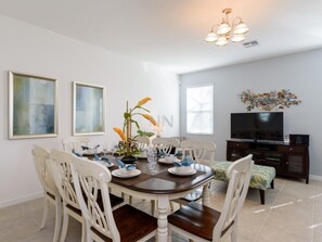 A dining area with a round table, white chairs, and a vase with flowers. There is a television and a cabinet in the background.