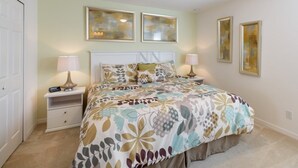 A bedroom with a large bed, two bedside tables, and a white headboard. There are two wall-mounted picture frames and a ceiling light.