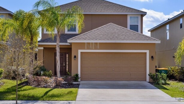 A two-story house with a brown garage door, a small front yard with a palm tree, and a clear blue sky.