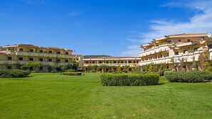 A hotel courtyard with neatly trimmed grass and hedges, featuring multi-story buildings with balconies and a clear blue sky. - Golfo Aranci - apartment for 2 people, directly on the beach, with terrace and sea-view (Golfo Aranci)