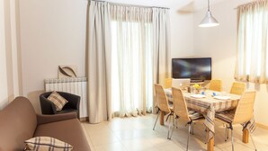 Una sala de estar moderna con un sofá, una mesa de comedor y sillas. Hay un televisor de pantalla plana montado en la pared, un radiador y una ventana con cortinas.