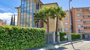 Un edificio moderno con ventanas amplias y una fachada curva, rodeado de un seto cuidado y palmeras.
