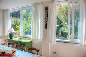 A dining area with a table set for two, a lamp, and a view of a garden through large windows.