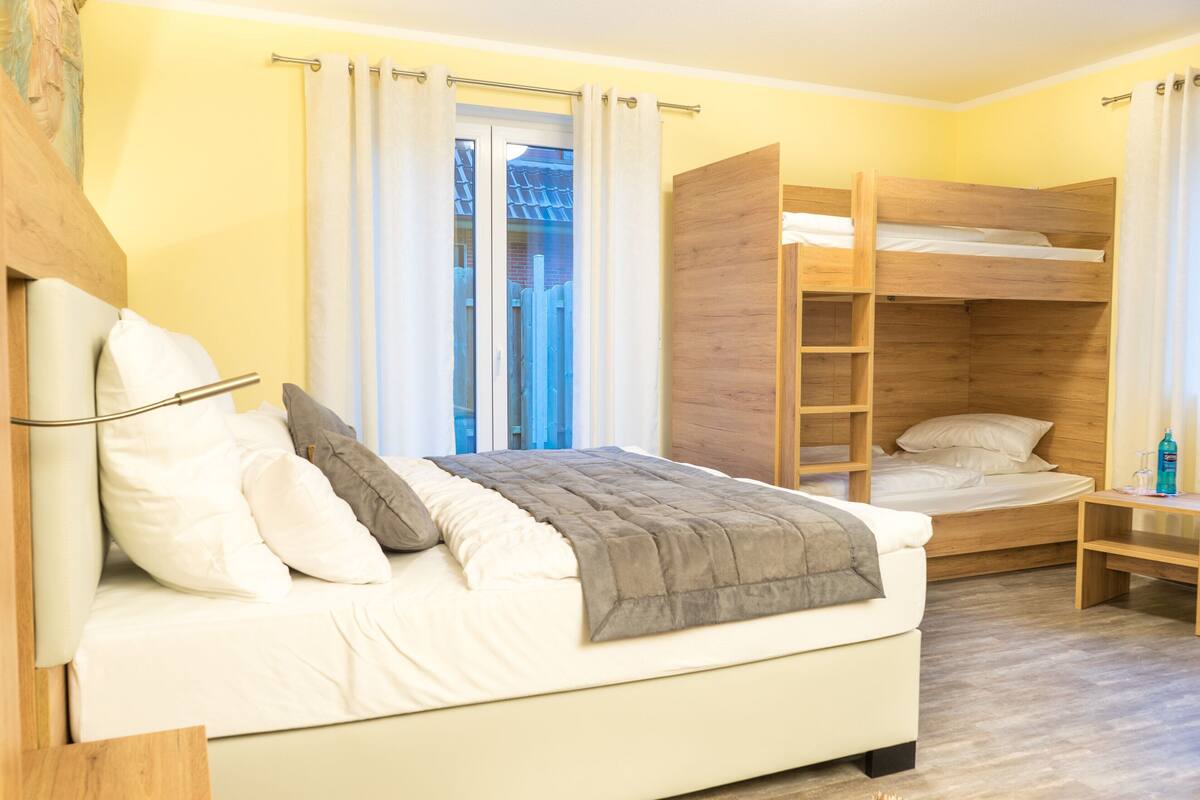 A room with a bunk bed, a single bed, a bedside table, and a window with curtains.