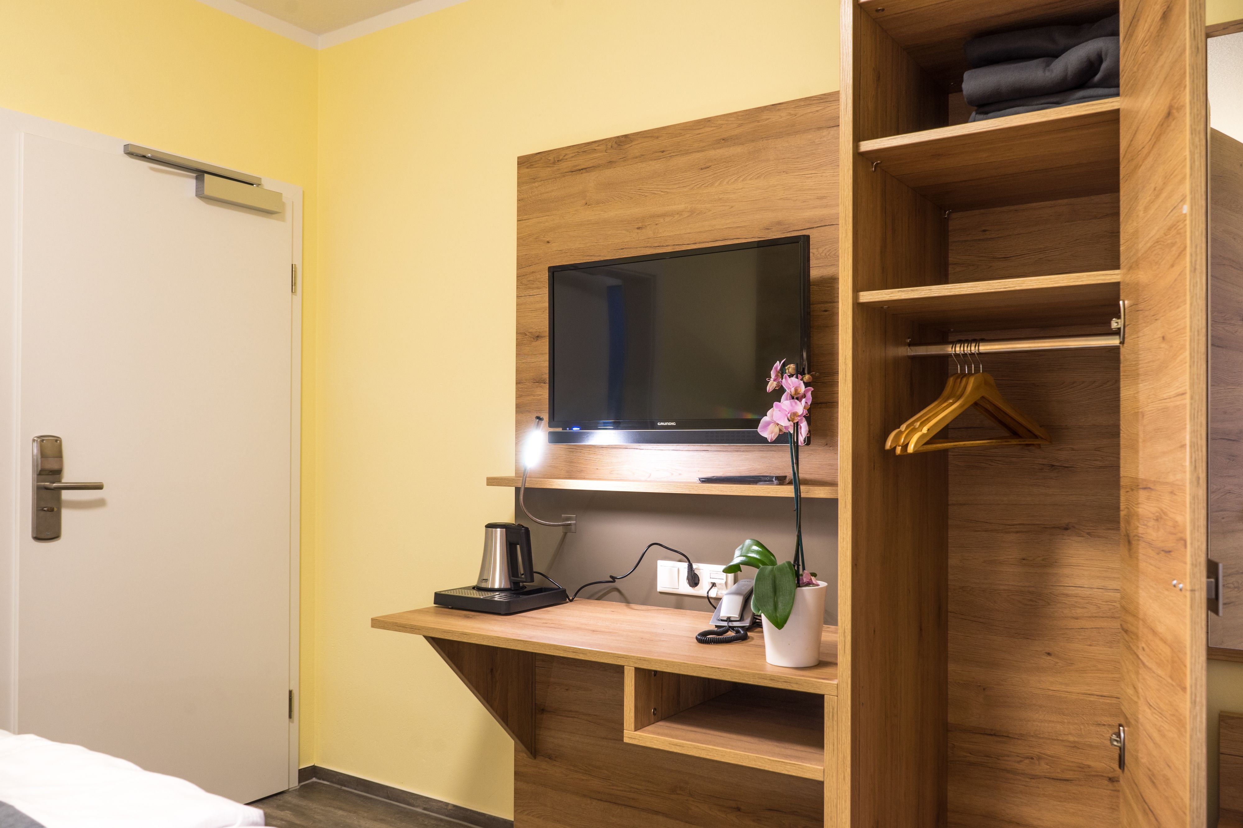 Une chambre d’hôtel avec un bureau en bois, une télévision, une machine à café, une plante et une armoire où sont suspendus des vêtements.