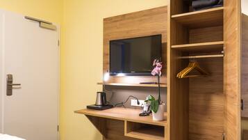 Une chambre d’hôtel avec un bureau en bois, une télévision, une machine à café, une plante et une armoire où sont suspendus des vêtements.
