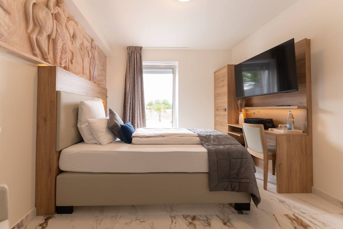 A modern hotel room with a bed, a desk, a flat-screen TV, and a large window with a view of greenery.