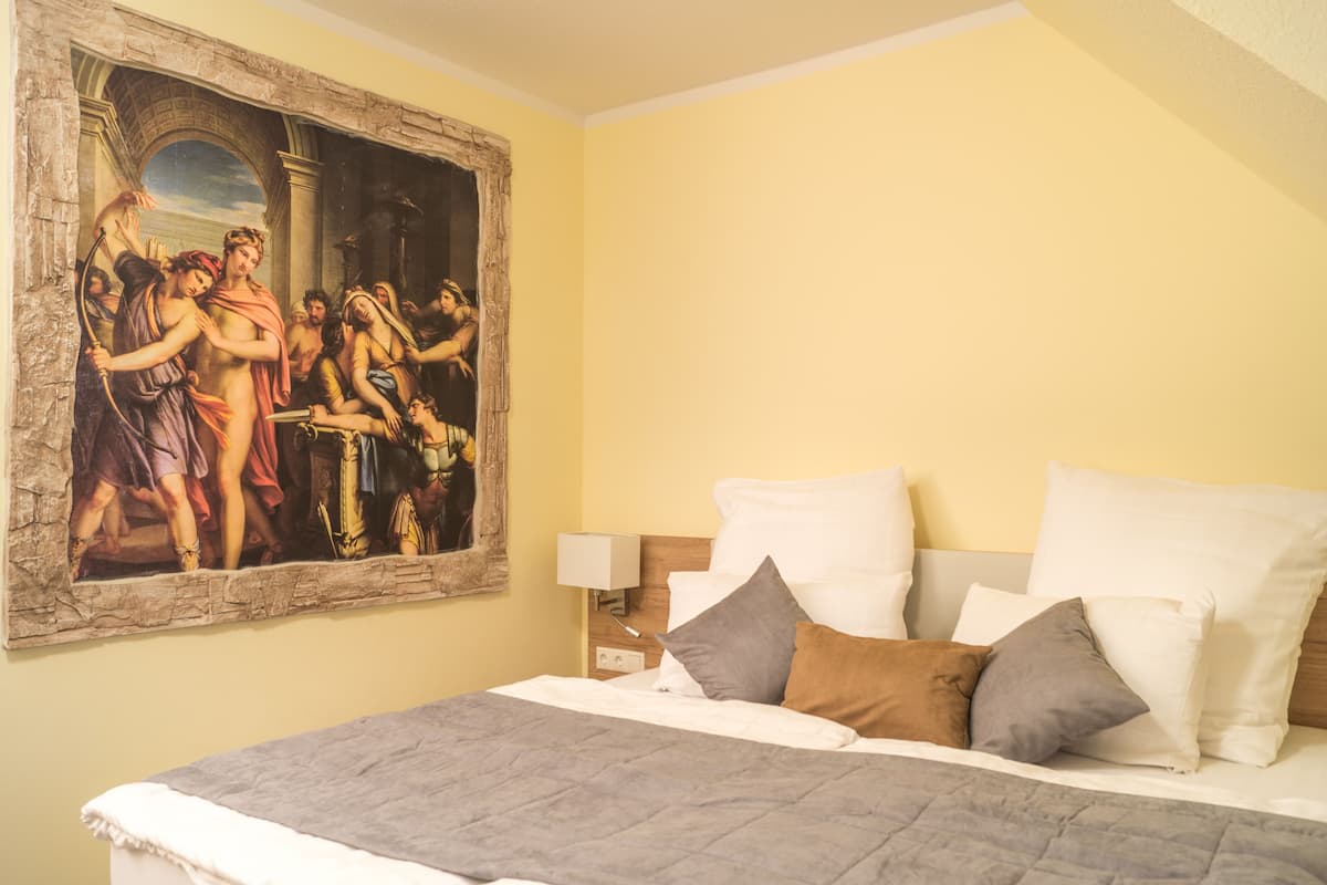 A hotel room with a large classical painting on the wall, a bed with white and gray bedding, and a nightstand with a lamp.