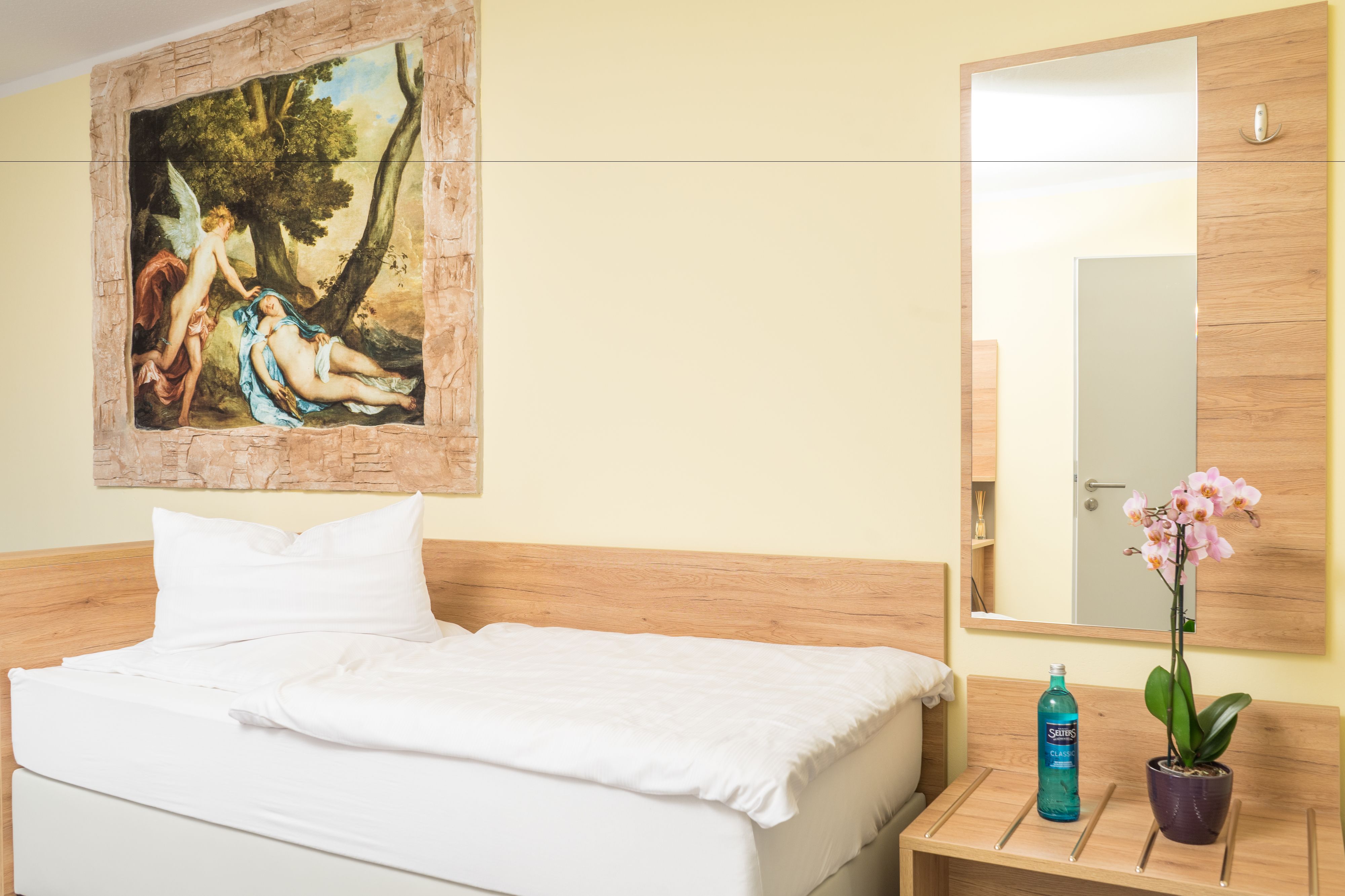 Una habitación de hotel con una cama, un cuadro, un espejo, una mesita de noche y una orquídea en maceta.